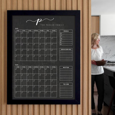 A wall-mounted family planner with calendar squares, menu, groceries, notes, and to-do sections. A woman stands in the background, holding papers in a modern kitchen.