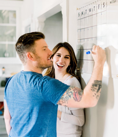 Close-up of a modern acrylic wall calendar with the word HAMILTON at the top. Days of the week, Monday to Saturday, are visible in bold black lettering. Gold mounting hardware is visible on the right side.