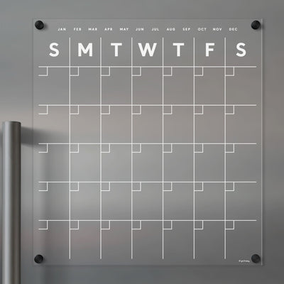 The Clear Acrylic Monthly Fridge Calendar by Girl Friday Shop features a frosted design for dry-erase markers, displaying a day grid labeled S M T W T F S. A sleek silver metallic cylinder enhances its modern look, perfect for organizing your month.