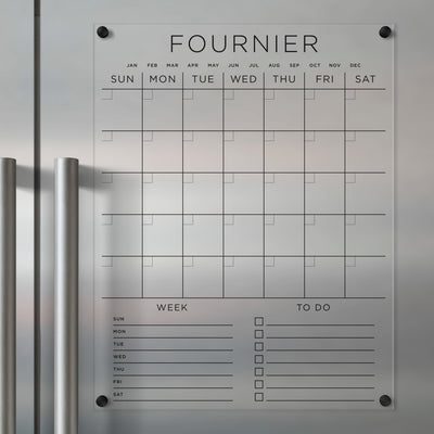 The Girl Friday Acrylic Fridge Calendar features custom family names, labeled days and months, weekly planning sections, and a to-do list. It mounts easily on stainless steel with vertical handles and is perfect for wet-erase markers.