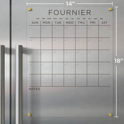 The Girl Friday Clear Acrylic Monthly Magnetic Fridge Calendar, customized with your family name, features a transparent grid for days and notes, supports wet-erase markers, has headers with days and months, gold-accented corners, and measures 14 x 18. Ideal for wall mounting.
