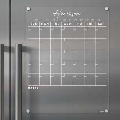 A Harrison clear acrylic calendar by Girl Friday is mounted on a dark surface with two handles. Its ideal for wet-erase markers and features days, months, and a bottom notes section.