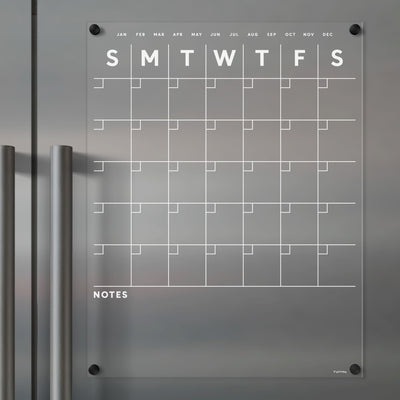 The Clear Acrylic Monthly Fridge Calendar by Girl Friday Shop elegantly adheres to stainless steel, featuring days of the week at the top and space for notes below. Its minimalist design includes wet-erase markers, with magnets securing each corner seamlessly.