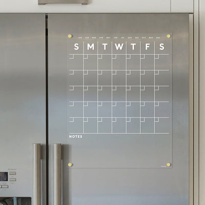 The Girl Friday Shops Clear Acrylic Monthly Magnetic Fridge Calendar is mounted on a metal refrigerator. It features an empty grid with white text, day spaces under Sunday to Saturday headings, and a Notes section at the bottom for use with a wet-erase marker.