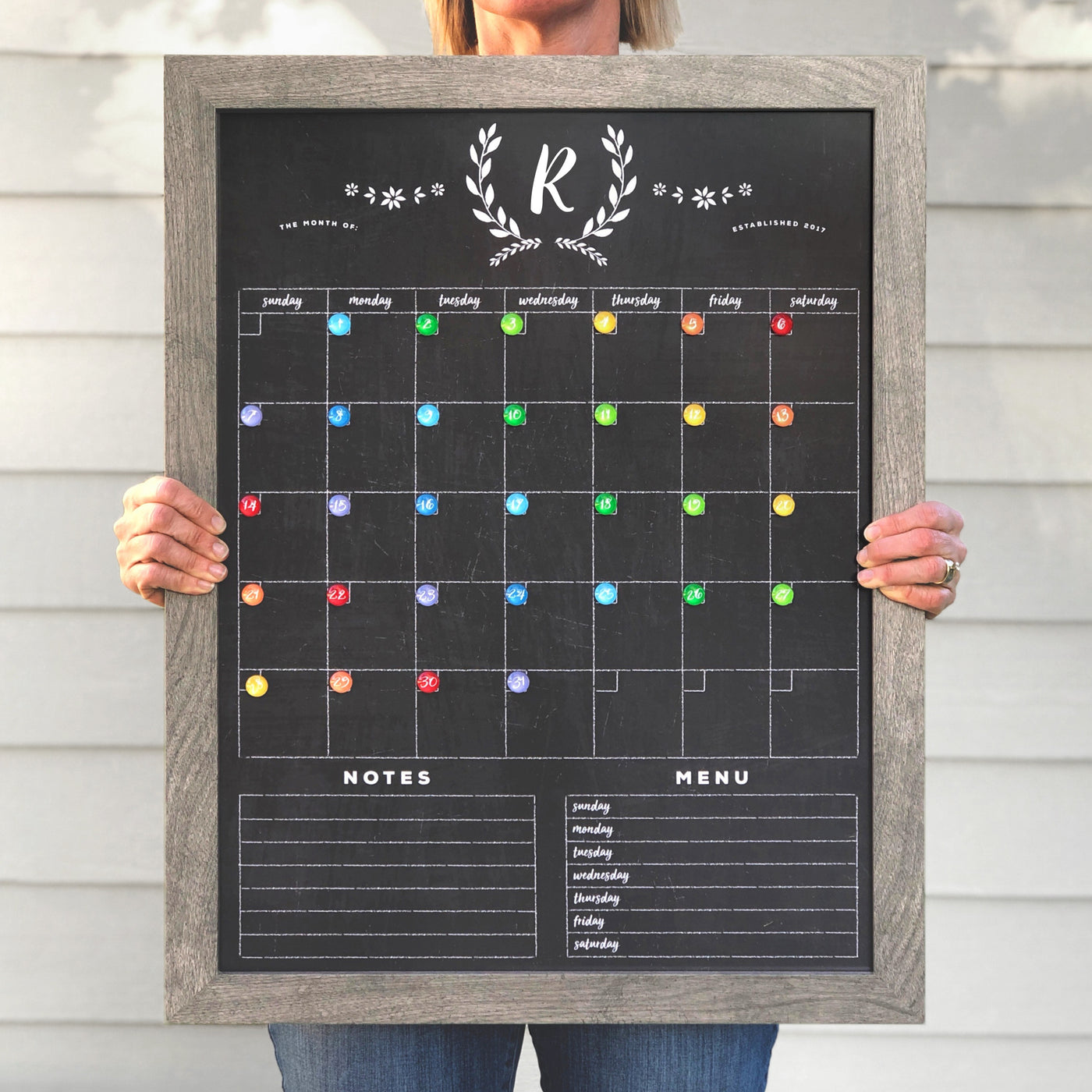 Someone holds the 2025 Personalized Dry Erase Framed Monthly 18 x 24 Calendar by CalendarGirlCompany. It features colorful magnets, a notes section, a menu plan at the bottom, and customizable elegant emblem with R at the top.