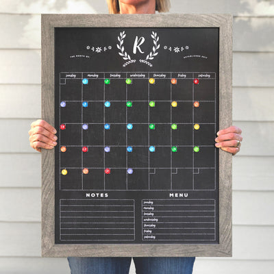 Someone holds the 2025 Personalized Dry Erase Framed Monthly 18 x 24 Calendar by CalendarGirlCompany. It features colorful magnets, a notes section, a menu plan at the bottom, and customizable elegant emblem with R at the top.