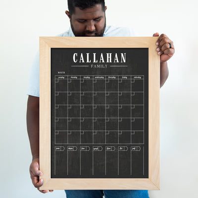 A person holds an 18x24 framed chalkboard calendar by CalendarGirlCompany, featuring CALLAHAN FAMILY at the top. It showcases spaces for days and months beautifully crafted with light wood. Ideal for using vibrant chalk ink markers to organize family plans.