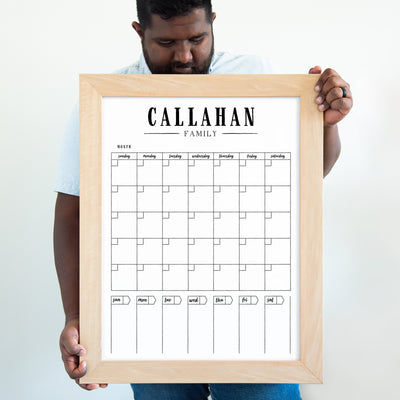 A person holds the CalendarGirlCompanys 18 X 24 framed whiteboard calendar (#18107) titled Callahan Family. It features a large acrylic surface with days of the week, empty daily squares, month indicators at the bottom, and is paired with a light wood frame ideal for chalk ink markers.