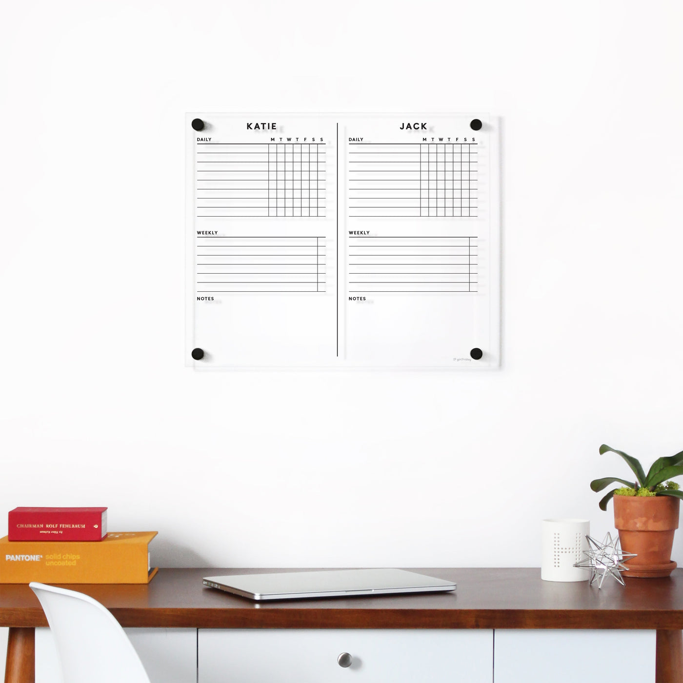 A neatly organized workspace features a wooden desk with a closed laptop, plant, candle, and stacked books. Above the desk hangs Girl Fridays Clear Acrylic Customizable Chore Chart for wall (1-3 children), divided into two columns labeled KATIE and JACK, perfect for jotting plans with wet-erase markers.