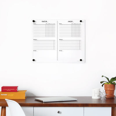 A neatly organized workspace features a wooden desk with a closed laptop, plant, candle, and stacked books. Above the desk hangs Girl Fridays Clear Acrylic Customizable Chore Chart for wall (1-3 children), divided into two columns labeled KATIE and JACK, perfect for jotting plans with wet-erase markers.