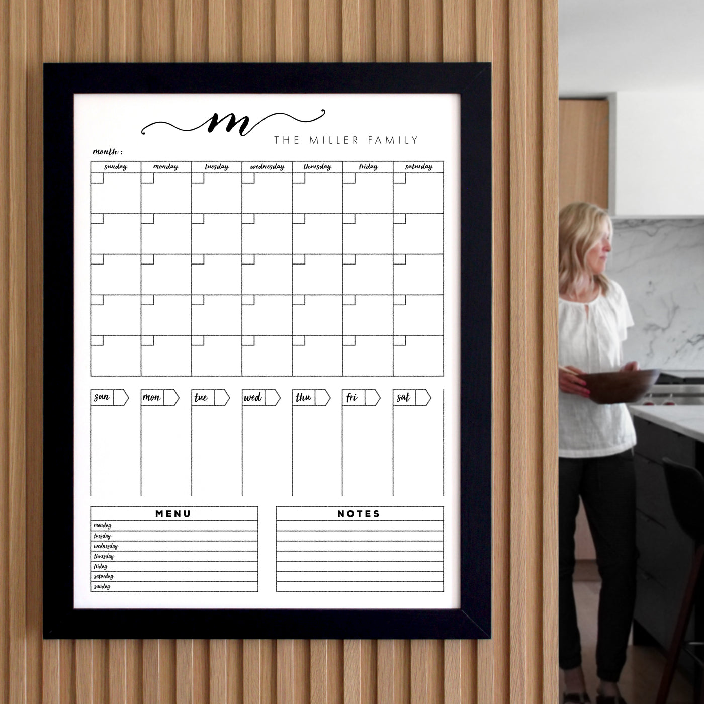 The 18 X 24 Framed Whiteboard Calendar by CalendarGirlCompany features a sleek black frame and an acrylic surface suitable for chalk ink markers. It displays a monthly calendar, menu, and notes section while someone stands in a kitchen holding a slightly blurred folder in the background.