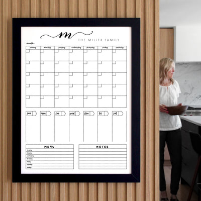 The 18 X 24 Framed Whiteboard Calendar by CalendarGirlCompany features a sleek black frame and an acrylic surface suitable for chalk ink markers. It displays a monthly calendar, menu, and notes section while someone stands in a kitchen holding a slightly blurred folder in the background.