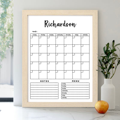 A Personalized Dry Erase Framed Monthly 18 x 24 Whiteboard Calendar (#18100) by CalendarGirlCompany rests on a shelf. Its premium acrylic surface includes sections for daily plans, notes, and a menu. An apple and small plant sit beside it as sunlight filters through the window casting gentle shadows.