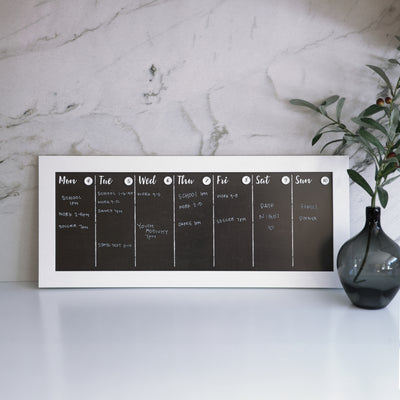 A 24 x 9 weekly chalkboard calendar from CalendarGirlCompany sits on a white desk, listing activities like school, soccer, youth activity, dance, and date night. A black vase with greenery is on the right against a marble wall—ideal for your favorite chalk ink markers.