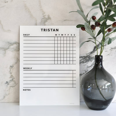 On a marble background, the White Acrylic Chore Chart by Girl Friday Shop showcases daily columns. The name Tristan is styled at the top. Nearby, a dark glass vase with green leaves and small berries enhances the scene. Its customizable with a premium dry erase surface, ideal for non-magnetic fridges.