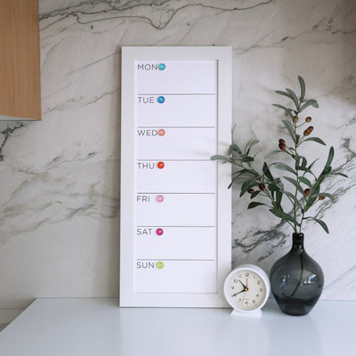 The CalendarGirlCompanys 9 x 24 weekly whiteboard calendar (#923) is mounted on a marble wall. It features a vertical Monday-to-Sunday schedule with colored circles for each day, complemented by a round clock and a vase with green branches on the premium acrylic surface.