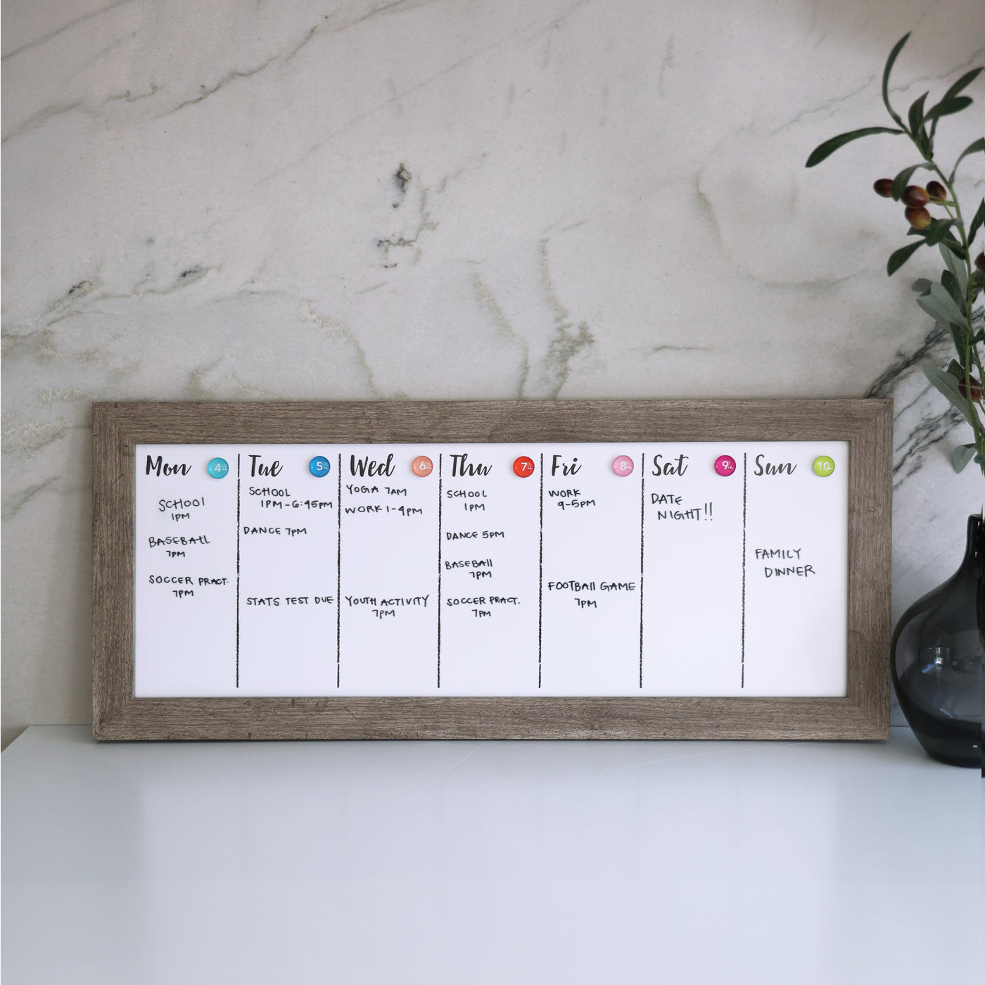The CalendarGirlCompanys 24 X 9 Framed Weekly Whiteboard Calendar (#922) with an acrylic surface sits elegantly on a white desk against a marble wall. Each days section lists activities like sports, work, and social events, while a small plant is partially visible on the desks right side.