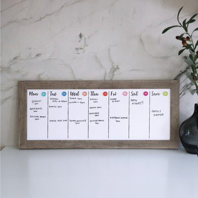 The CalendarGirlCompanys 24 X 9 Framed Weekly Whiteboard Calendar (#922) with an acrylic surface sits elegantly on a white desk against a marble wall. Each days section lists activities like sports, work, and social events, while a small plant is partially visible on the desks right side.