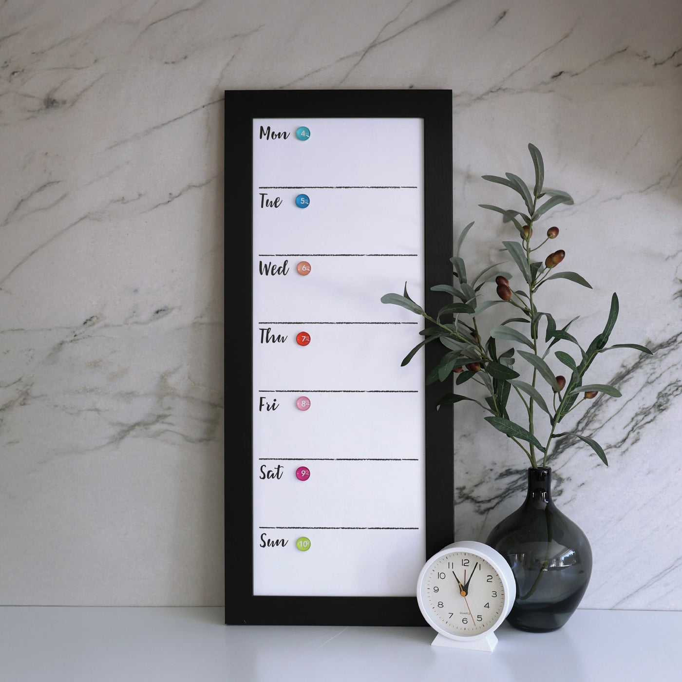 A 9 X 24 weekly whiteboard calendar by CalendarGirlCompany rests on a marble surface, with days listed vertically and highlighted by colored chalk dots. Nearby, a dark glass vase with green branches and a white clock add elegance.