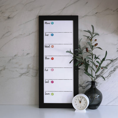 A 9 X 24 weekly whiteboard calendar by CalendarGirlCompany rests on a marble surface, with days listed vertically and highlighted by colored chalk dots. Nearby, a dark glass vase with green branches and a white clock add elegance.
