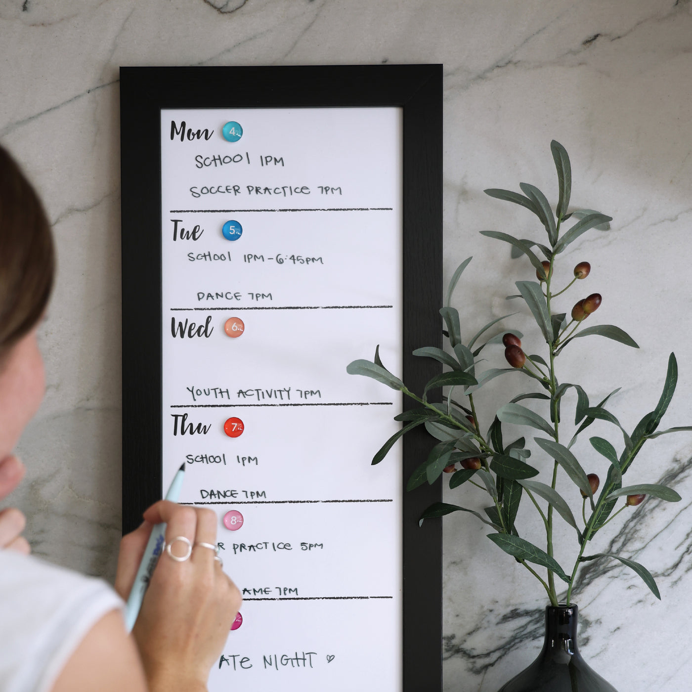 A person uses chalk ink markers on a CalendarGirlCompany 9 X 24 weekly whiteboard calendar (#921) hanging on a marble wall, listing activities like school, soccer practice, and dance. Nearby is an olive branch in a black vase.
