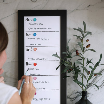 A person uses chalk ink markers on a CalendarGirlCompany 9 X 24 weekly whiteboard calendar (#921) hanging on a marble wall, listing activities like school, soccer practice, and dance. Nearby is an olive branch in a black vase.