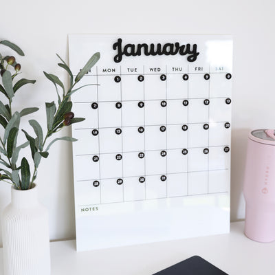 A White Acrylic Monthly Calendar by Girl Friday Shop, featuring a sleek black and white design with numbered circles for January from Sunday to Saturday and a dry erase surface, sits stylishly on a desk alongside a white vase with green leaves and a pink insulated tumbler.
