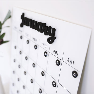 The White Acrylic Monthly Calendar by Girl Friday Shop showcases January in bold black letters. It starts the week with Sunday, includes a small plant design, and can be easily mounted using Command Strips for a sleek workspace display.