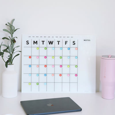 The White Acrylic Monthly Calendar by Girl Friday Shop, featuring a dry erase surface and a notes section on the side, displays colorful stickers marking days. It sits on a white desk alongside a closed laptop, white vase with greenery, and pink water bottle. This calendar requires no mounting.
