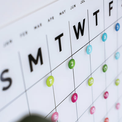 A close-up of the White Acrylic Monthly Calendar by Girl Friday Shop shows days from Sunday, with multicolored buttons marking dates in early June on its dry erase surface against a white background.