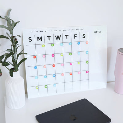 The desk showcases a Girl Friday Shop White Acrylic Monthly Calendar with weekdays at the top and colored circular markers. A potted plant sits on the left next to a closed laptop, while a pink cylinder is mounted on the right using command strips.