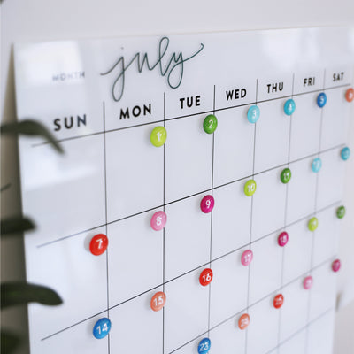 The Girl Friday Shops White Acrylic Monthly Calendar for July displays Sunday to Saturday. Colorful magnetic markers add vibrancy, with a green plant on the left. Suitable for non-magnetic fridges, it offers an easy-to-use dry erase surface for seamless planning.