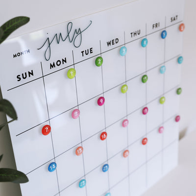The White Acrylic Monthly Calendar by Girl Friday Shop hangs on the wall, offering a dry erase surface. Colorful blue, pink, and green pins mark each day. A leafy green plant adds a refreshing touch to your modern scheduling tool.