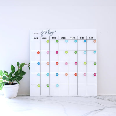 The White Acrylic Monthly Calendar by Girl Friday Shop features colorful circular date markers and a dry erase surface for easy updates. A small potted plant with green leaves on a marble surface adds brightness, ideal for non-magnetic fridges or offices.