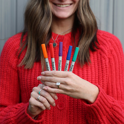 A woman in a red sweater holds a Girl Friday Shops FrauLiner wet-erase marker set—fine tip—with colorful, non-toxic caps in orange, red, pink, purple, blue, and green. Smiling as her light pink nails catch the light, shes ready to create vibrant designs on acrylic boards.