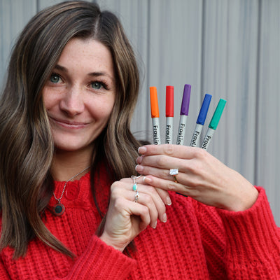 A person with long hair in a red sweater holds the FrauLiner wet-erase marker set by Girl Friday Shop, featuring vibrant colors like orange, red, purple, blue, and green. They pose confidently against a gray corrugated backdrop, ready to create on acrylic boards using the fine tip markers.