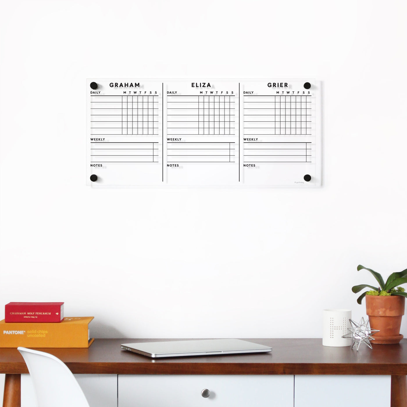 A Girl Friday Clear Acrylic Customizable Chore Chart for 1-3 children hangs on the wall, divided into sections for Graham, Eliza, and Grier with wet-erase markers. Below, a wooden desk holds stacked books, a laptop, a potted plant, and a pen holder.