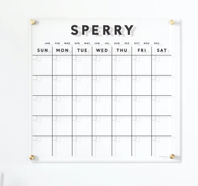 The Girl Friday Clear Acrylic Customizable Monthly Wall Calendar is a minimalist white calendar with a grid format displaying January to December and days Sunday to Saturday; Sperry is printed on top, and its compatible with wet-erase markers for easy updates.