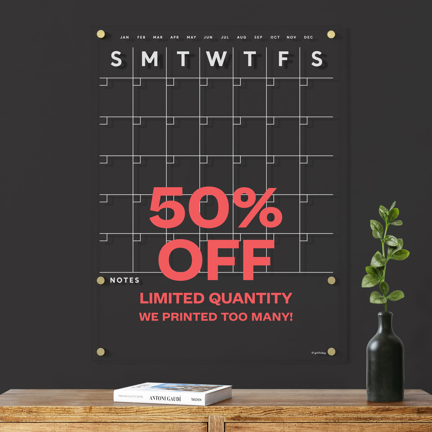 The Large Acrylic Calendar with Bottom Notes by Girl Friday Shop boasts a spacious monthly grid, weekdays in white text, and is ideal for wet-erase markers. It’s available at 50% off due to overstock. Hurry, limited supply! A small plant and wooden table enhance its sleek setting.