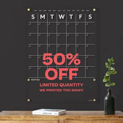 The Large Acrylic Calendar with Bottom Notes by Girl Friday Shop boasts a spacious monthly grid, weekdays in white text, and is ideal for wet-erase markers. It’s available at 50% off due to overstock. Hurry, limited supply! A small plant and wooden table enhance its sleek setting.