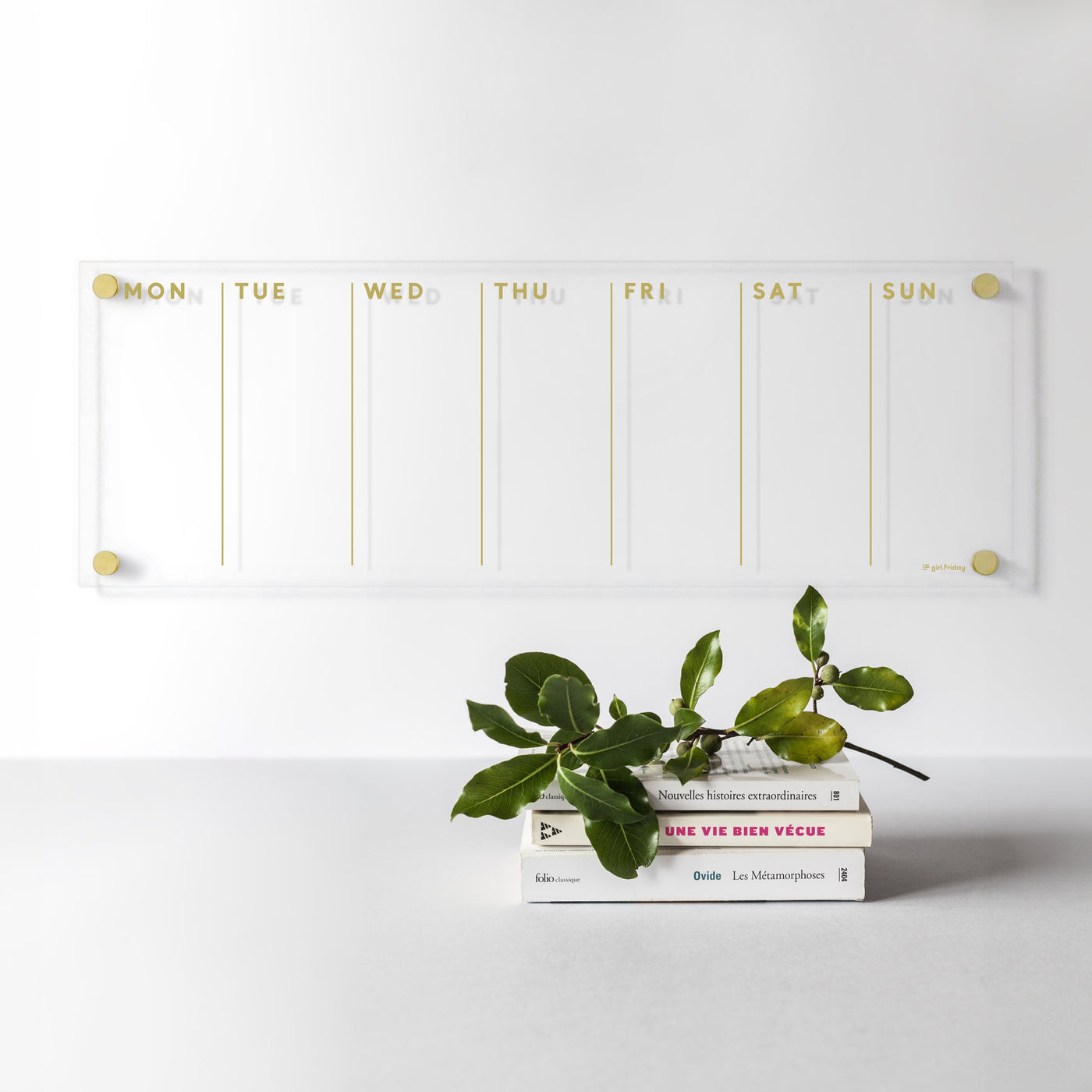 The Girl Friday Shops Acrylic Calendar showcases a chic weekly layout with gold text from Monday to Sunday. Three stacked books and leafy greens complement the minimalist style, ideal for planning with wet-erase markers on a light gray surface against a white wall.