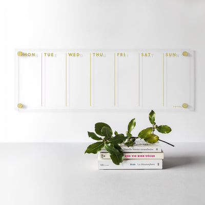The Girl Friday Shops Acrylic Calendar showcases a chic weekly layout with gold text from Monday to Sunday. Three stacked books and leafy greens complement the minimalist style, ideal for planning with wet-erase markers on a light gray surface against a white wall.