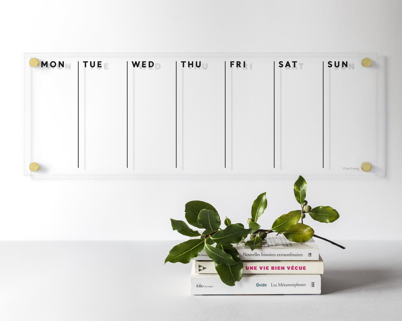 The Vertical Clear Acrylic Weekly Wall Calendar by Girl Friday Shop adorns the white wall, showcasing weekday headers. Below, a stack of three books and a small green-leafed branch complement wet-erase markers, all set against a neutral-toned minimalist background.