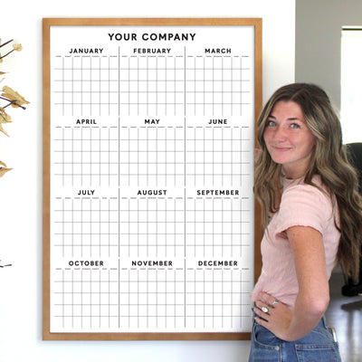A woman with long hair in a pink shirt stands beside a 2025 Girl Friday Vertical Yearly Calendar titled Your Company, featuring monthly rows. She smiles, hand on hip, with a yellow-flowered plant visible on the left.
