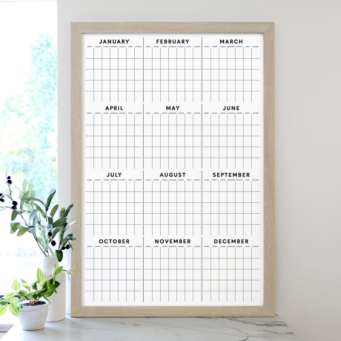 The 2025 Framed Yearly Whiteboard Calendar by Girl Friday features a minimalist design framed in wood, with a premium acrylic surface displaying all months from January to December. Ideal for use with wet-erase markers, its complemented by a small plant sitting nearby on the white surface.