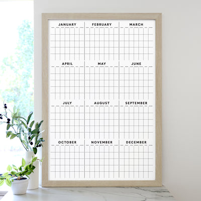 The 2025 Framed Yearly Whiteboard Calendar by Girl Friday features a minimalist design framed in wood, with a premium acrylic surface displaying all months from January to December. Ideal for use with wet-erase markers, its complemented by a small plant sitting nearby on the white surface.