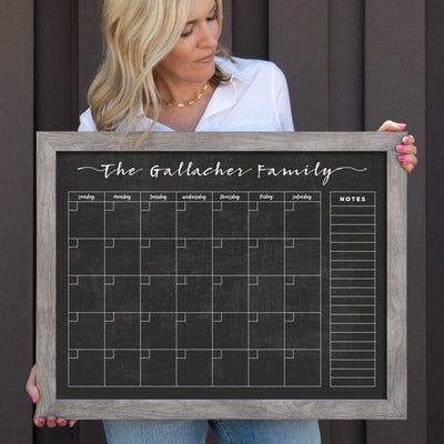 A woman in a white shirt and jeans holds the CalendarGirlCompanys 24 X 18 Framed Chalkboard Calendar #18203, titled The Gallacher Family. Featuring vibrant chalk ink marker colors, it has a Sunday-to-Saturday weekly grid and notes section on the right, set against a dark background.
