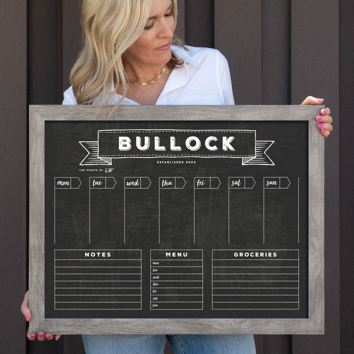 Someone holds a 24 X 18 Framed Chalkboard Calendar (#18210) by CalendarGirlCompany, featuring sections for each day, notes, menu, and grocery list. The customizable frame displays BULLOCK and an established date, allowing for personal creativity using colorful chalk ink markers.