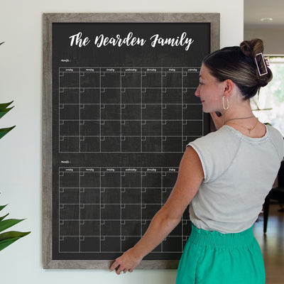 A woman with a bun hairstyle and wearing a light top and green pants stands next to the 24 x 36 Framed Chalkboard Calendar by CalendarGirlCompany. Shes adjusting the customizable frame on a white wall, with chalk ink markers nearby for updates. A potted plant is visible on the left.