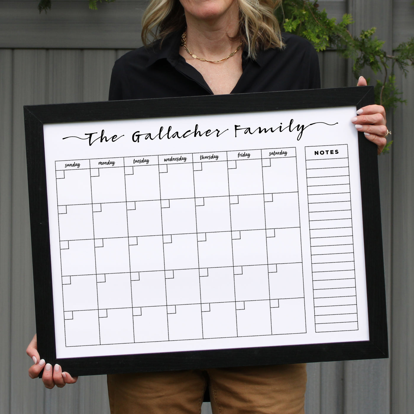 A person confidently stands against a gray wall holding the 24 X 18 Framed Whiteboard Calendar (#18203) by CalendarGirlCompany, labeled The Gallacher Family. The calendar includes a weekly grid and notes section, ideal for chalk ink markers. The individual is dressed in a black top and beige pants.